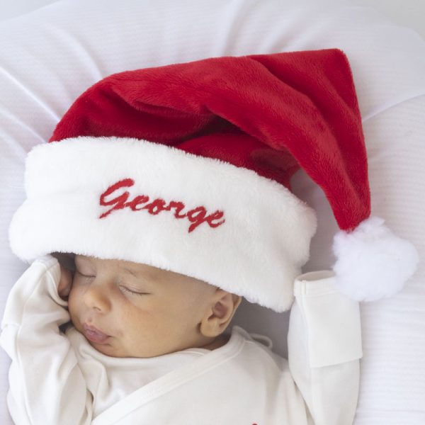 A baby wearing a personalised santa hat with the name George embroidered in red thread on the edge.