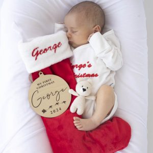 A baby wearing a personalised long-sleeve romper with a Christmas stocking, wooden ornament and bear rattle personalised gift.