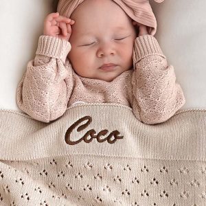 A baby laying under a Personalised Beige Pointelle Knitted Baby Blanket with the name Coco.