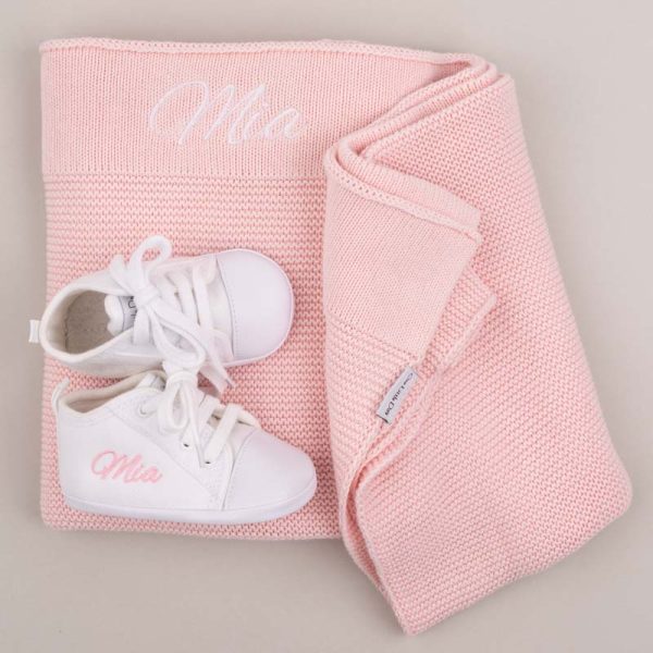 White Baby Shoes in front of a Pink Knitted Blanket and both embroidered with the name Mia.