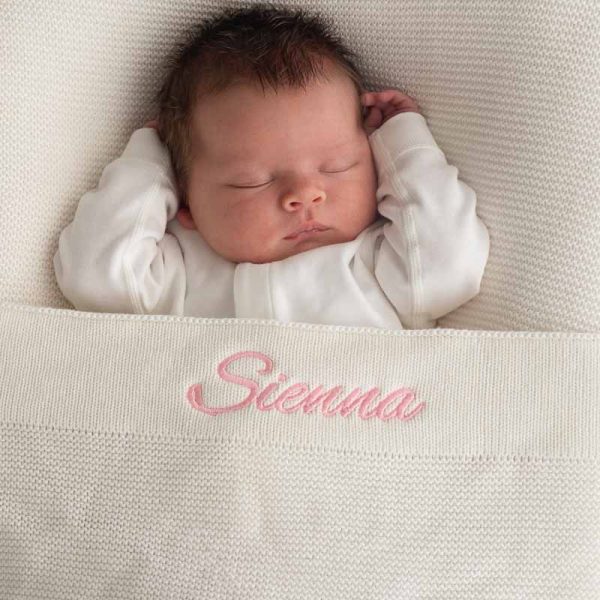 Baby laying under a white knitted cotton blanket personalised with the name Sienna with pink thread..