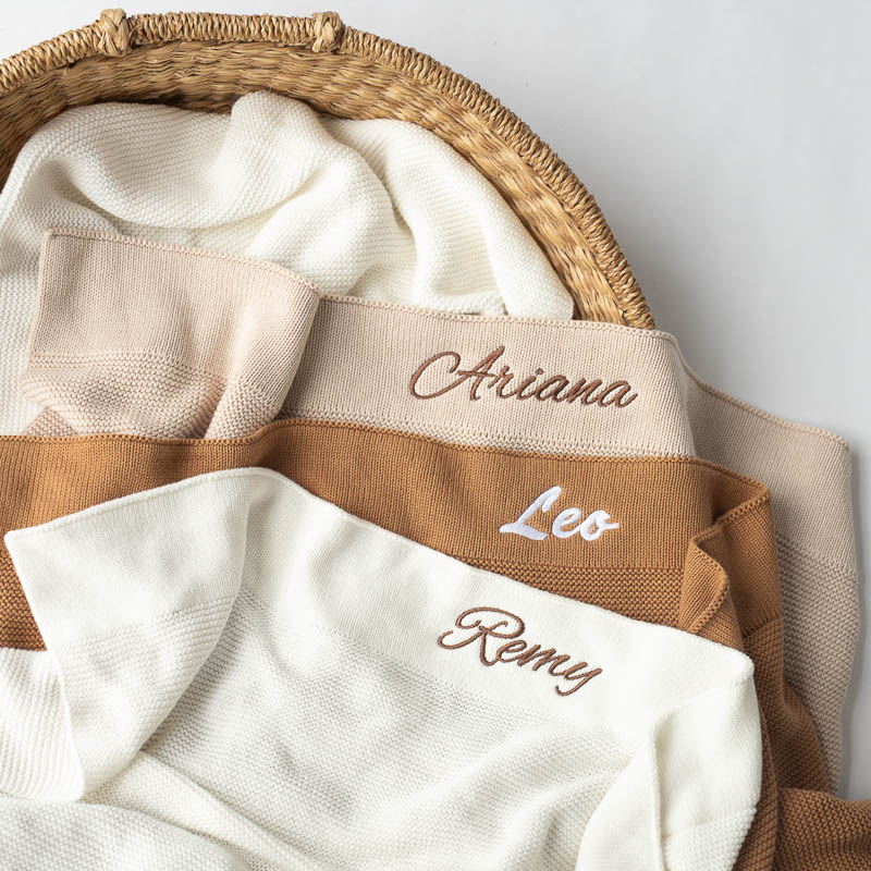 Personalised beige, brown and white knitted baby blankets with name embroidery laid out in a basket.