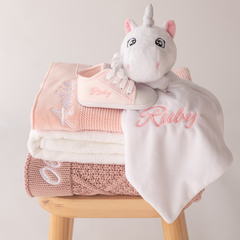 A white unicorn baby comforter sitting on top of 3 stacked personalised knitted blankets.