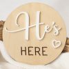 A birth wood birth announcement disc that says "He's Here" with a heart in white acrylic overlayed.