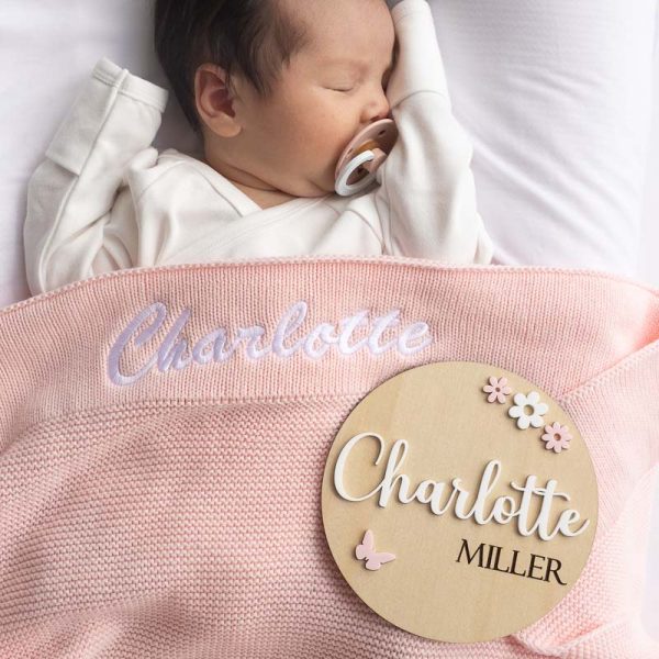 A baby laying under a pink personalised baby blanket and a birth flowers & butterfly birth announcement disc.